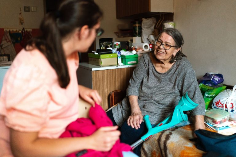 Pflegekraft unterstützt eine Seniorin bei hauswirtschaftlichen Aufgaben im Wohnzimmer