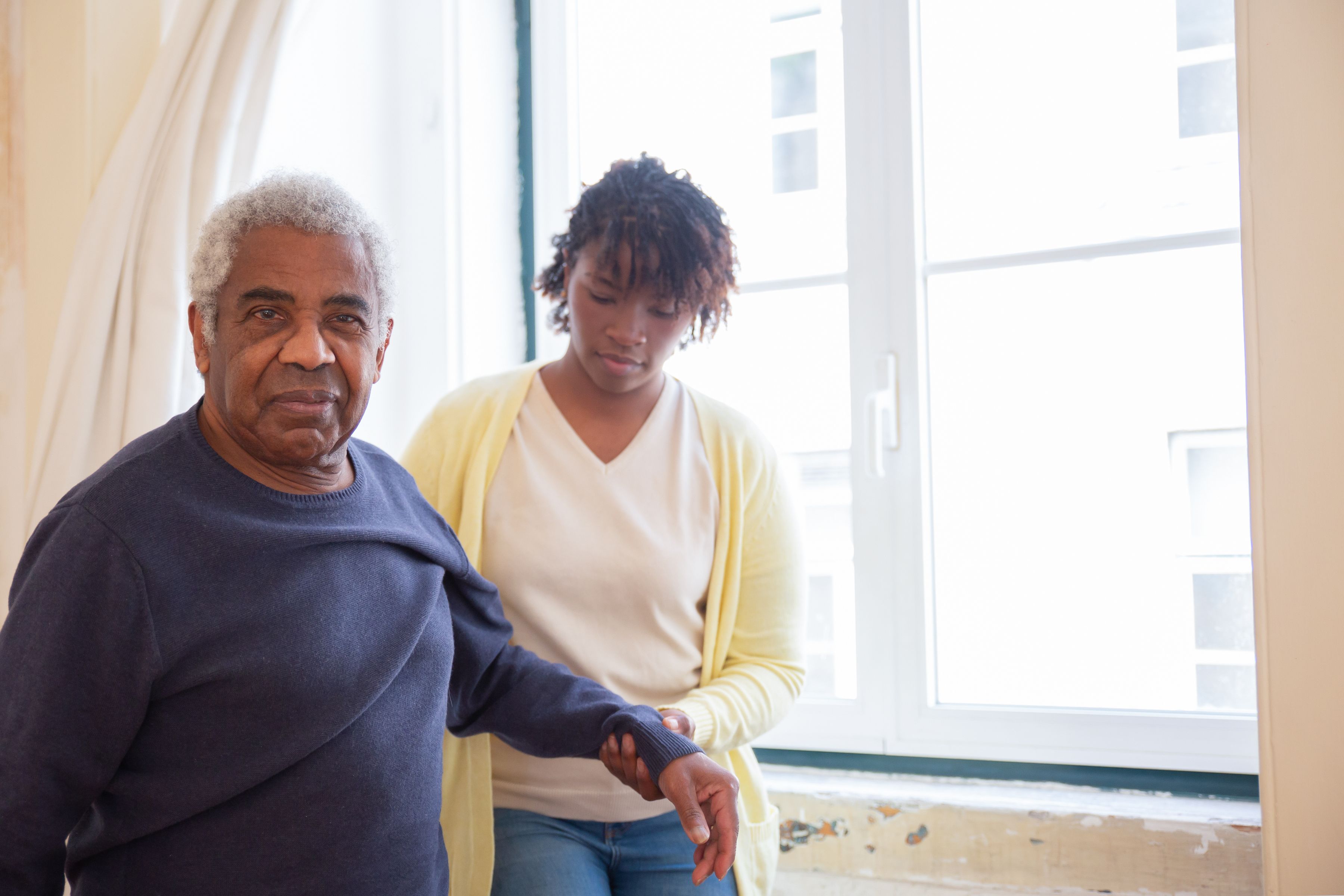 Pflegekraft begleitet einen Senior Arm in Arm durch die Wohnung